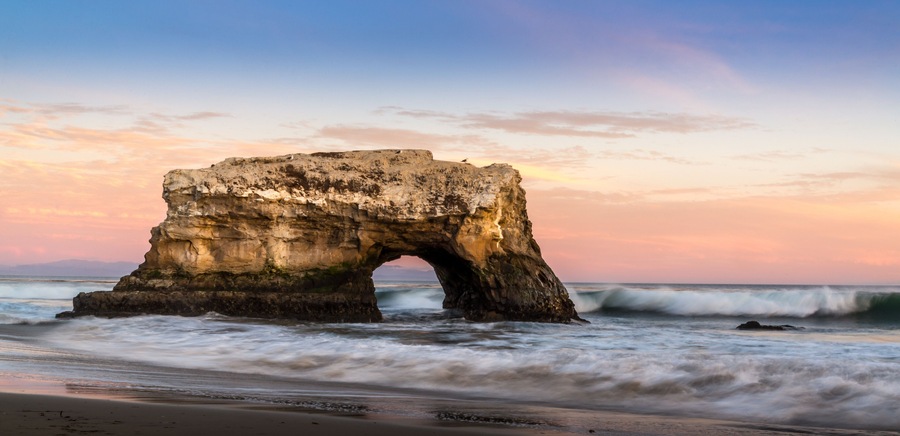 A natural rock arch formation on a beach with waves crashing below.