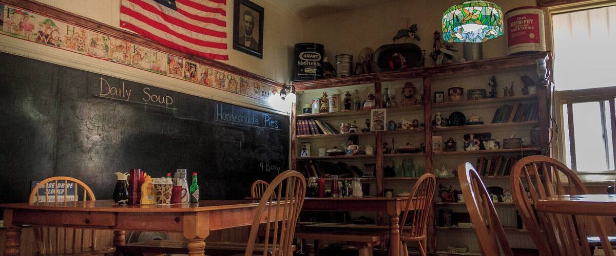 A restaurant interior with wooden furniture, an American flag, a chalkboard menu, and a bookshelf.