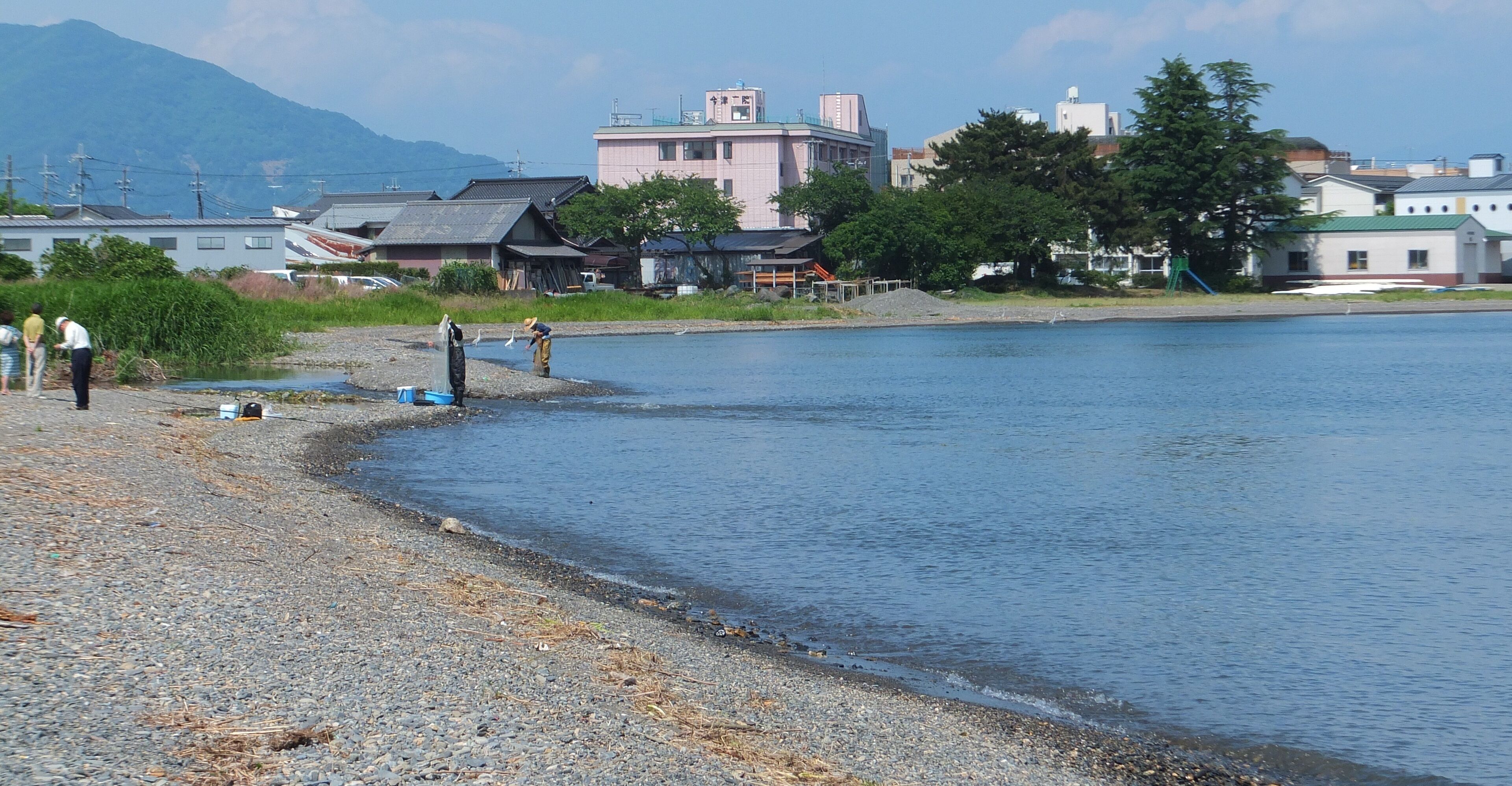 A pebbly beach with people fishing, a calm lake, and buildings in the background.