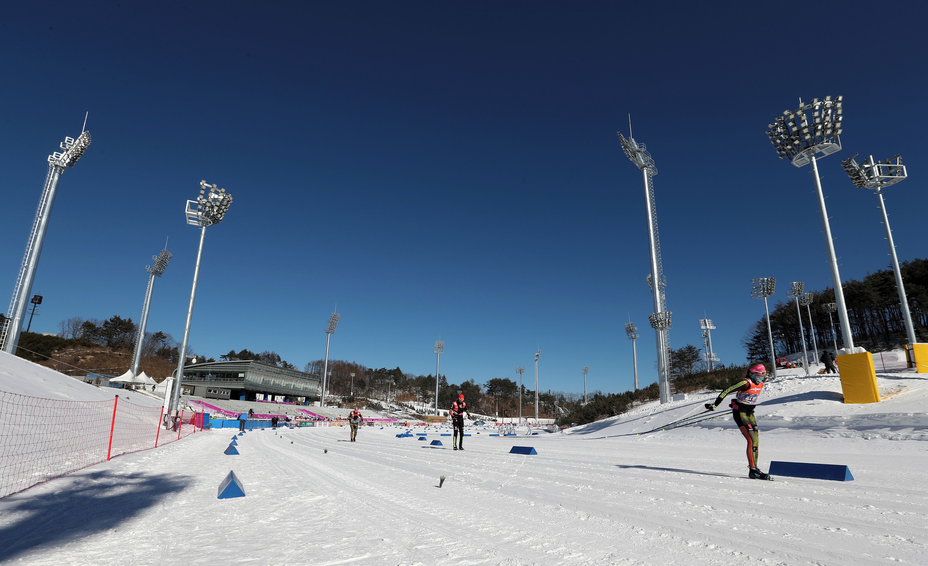 A snowy cross-country skiing course with multiple lanes, light poles, and a clear blue sky.