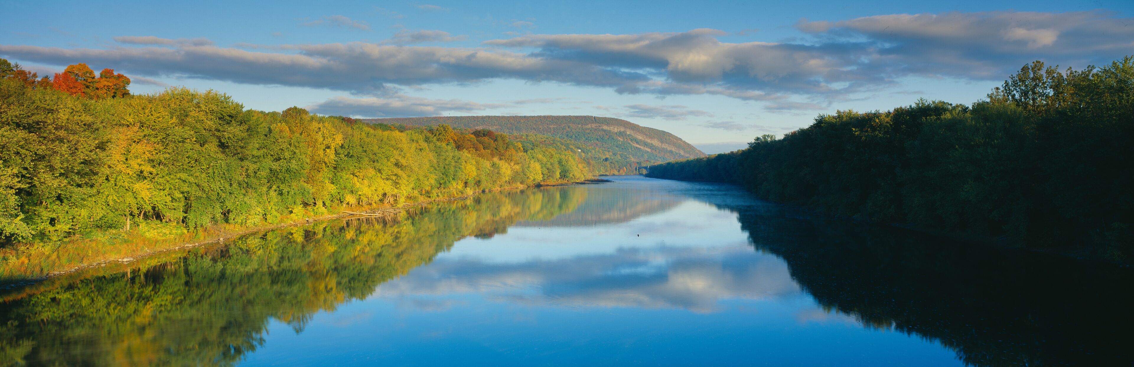 A calm river reflecting the surrounding autumn foliage and a distant hill.