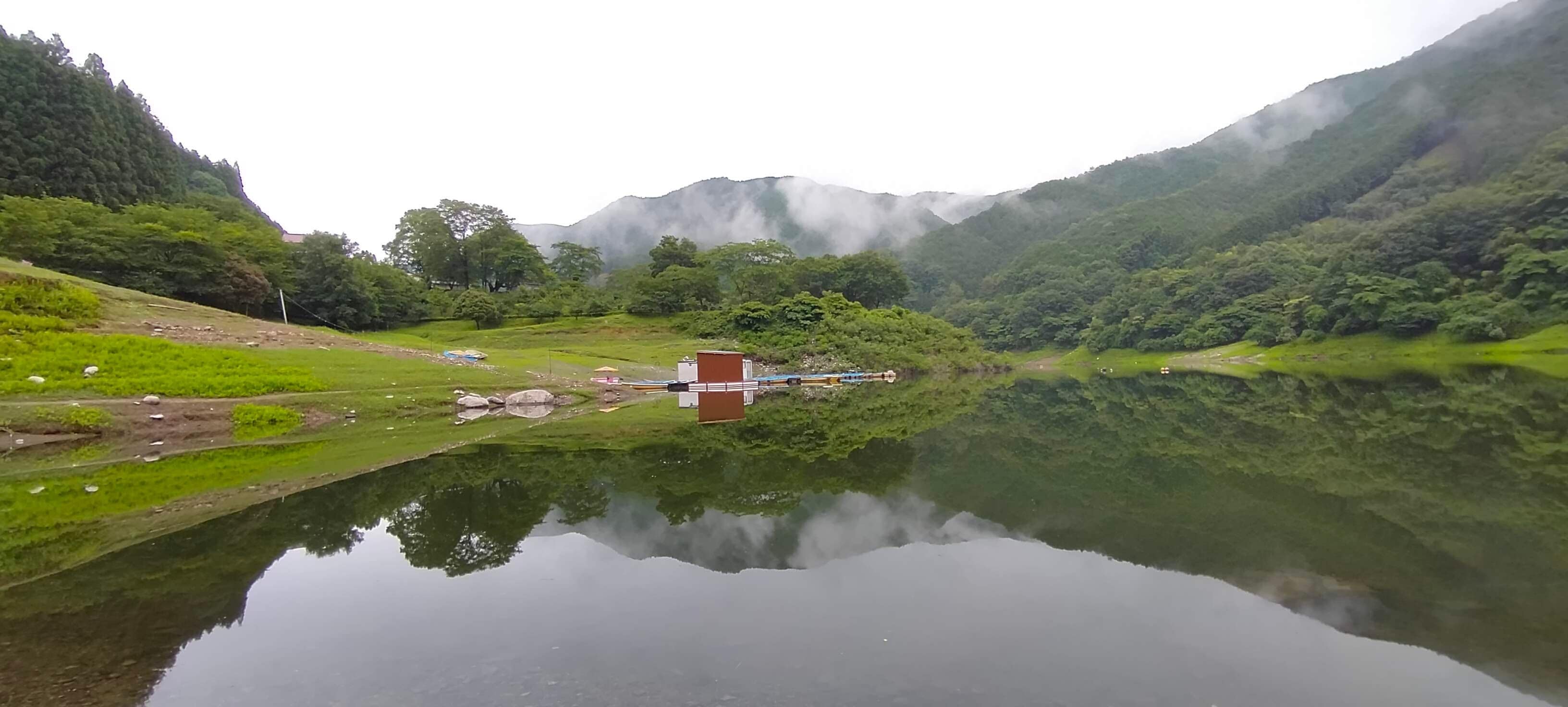 A calm lake with a small building and a train in the distance, surrounded by lush green hills and mist.