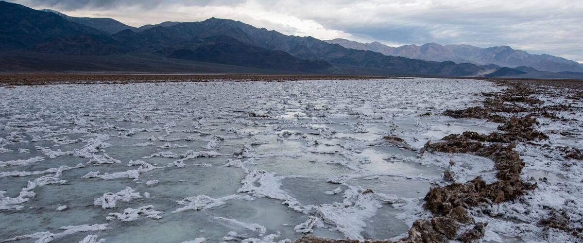 A vast salt flat with white salt crusts and a distant mountain range under a cloudy sky.