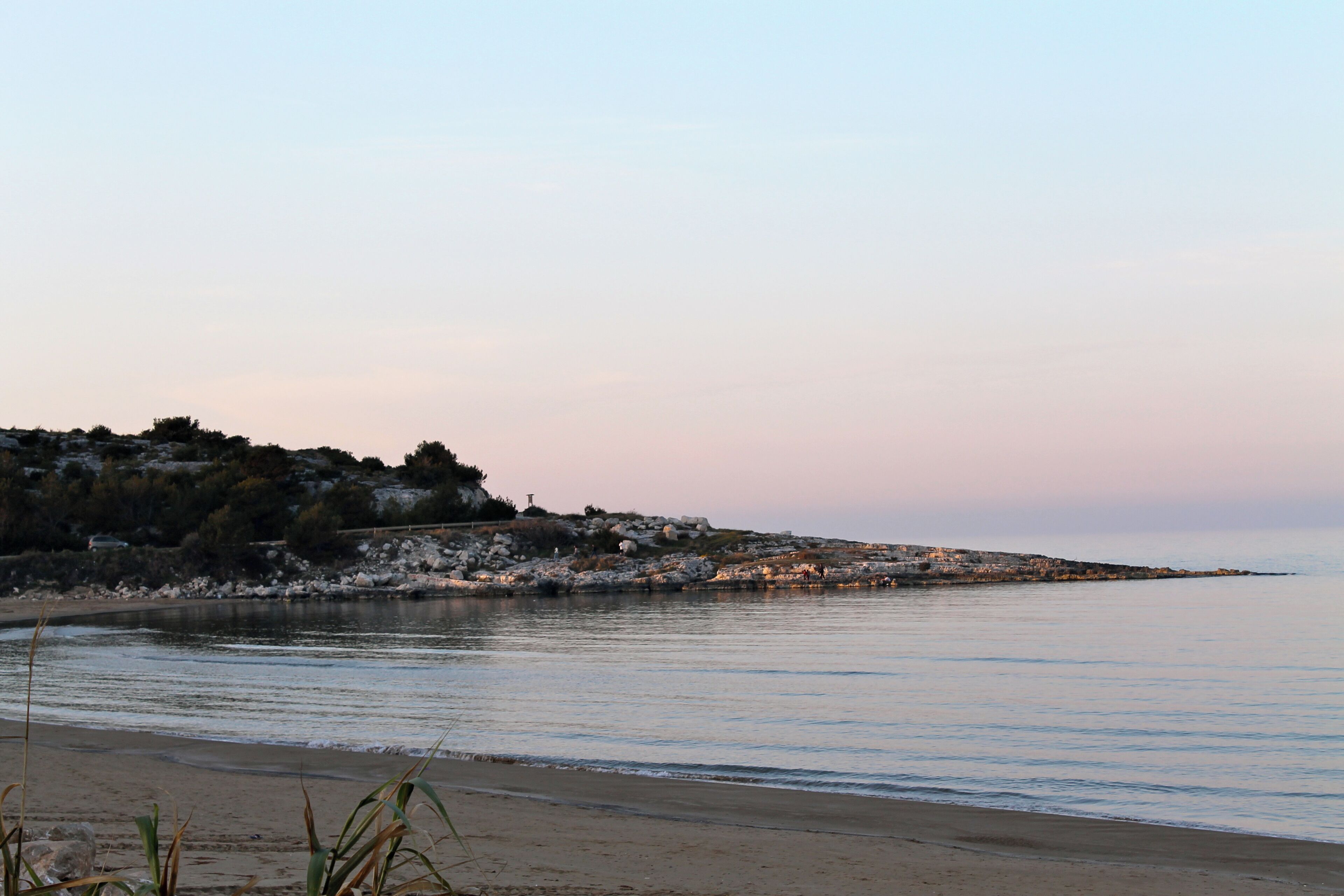 A coastal landscape with a sandy beach, calm water, rocky outcrops, and a hill with sparse vegetation.