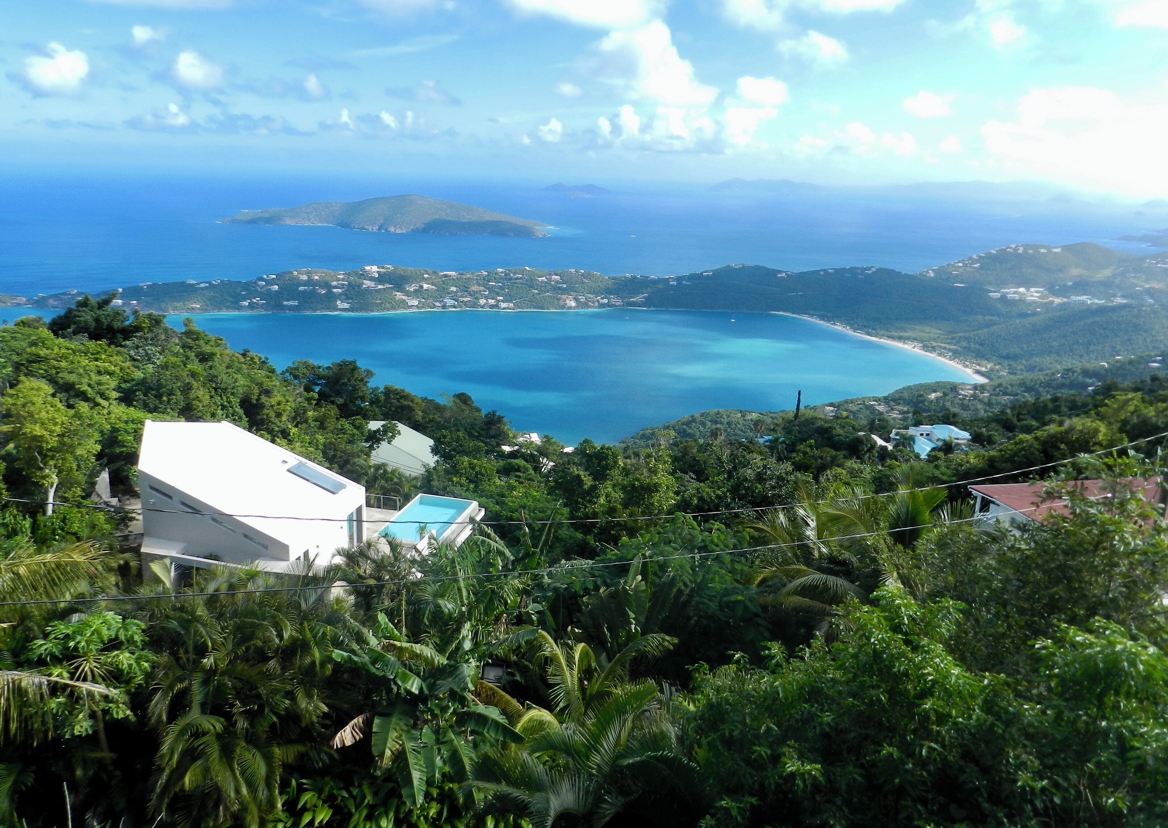 A coastal landscape with a clear blue sea, lush greenery, and a white building with a pool.