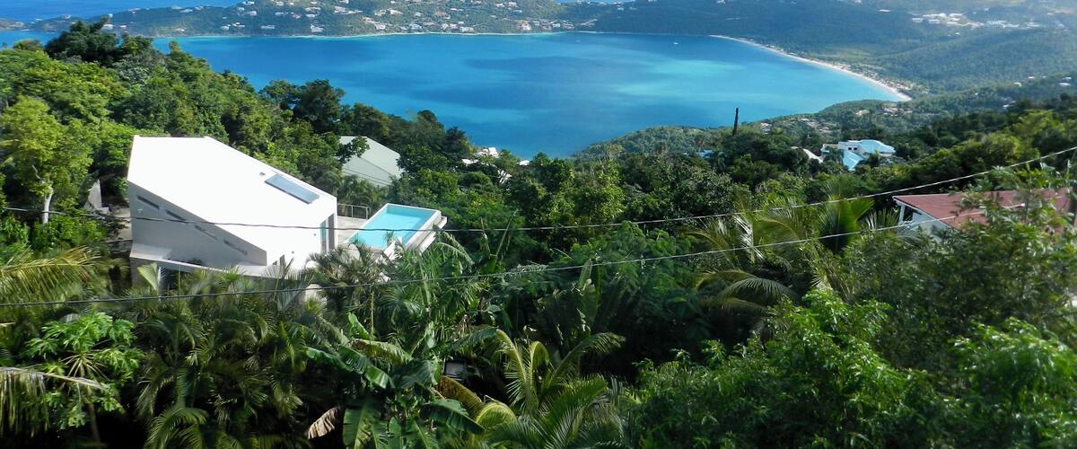 A coastal landscape with a clear blue sea, lush greenery, and a white building with a pool.