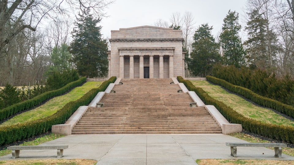 A grand building with a central staircase leading to a large entrance, surrounded by neatly trimmed hedges and trees.