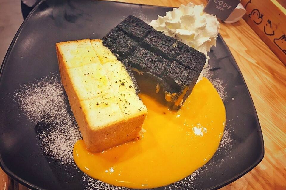 A dessert with a square piece of bread and a square piece of chocolate on a black plate, with a yellow sauce and whipped cream.