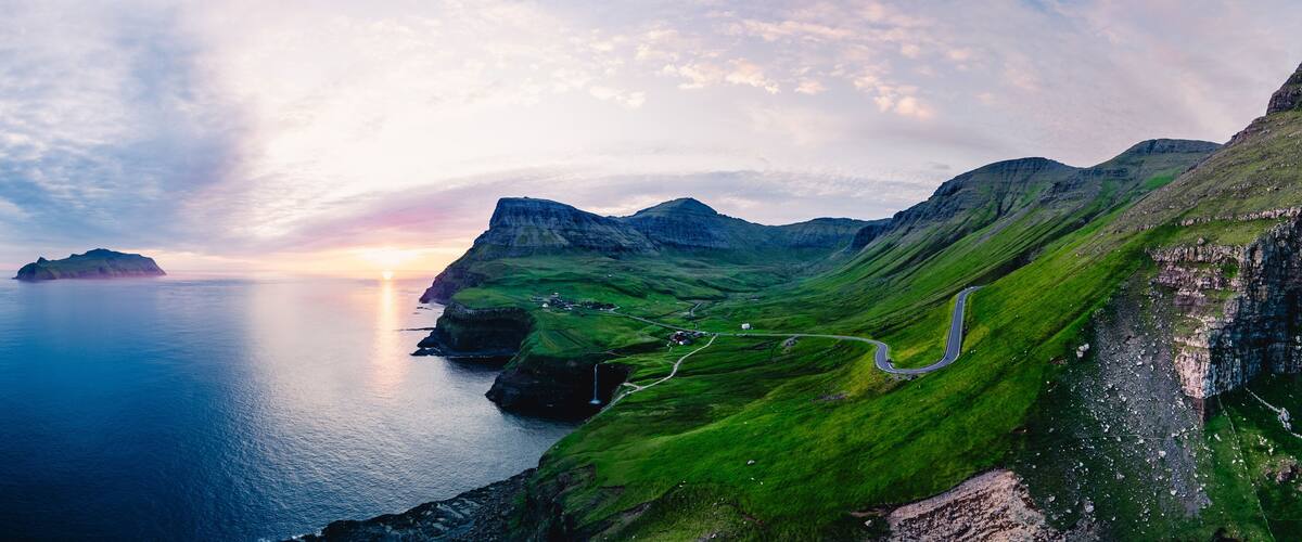 A coastal landscape with green hills, a winding road, and a body of water at sunset.