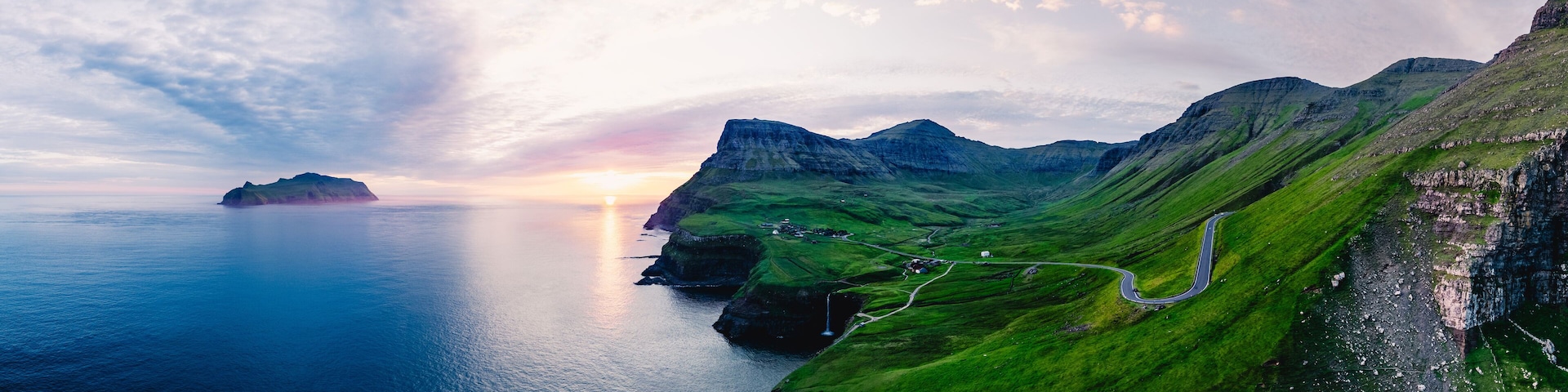 A coastal landscape with green hills, a winding road, and a body of water at sunset.