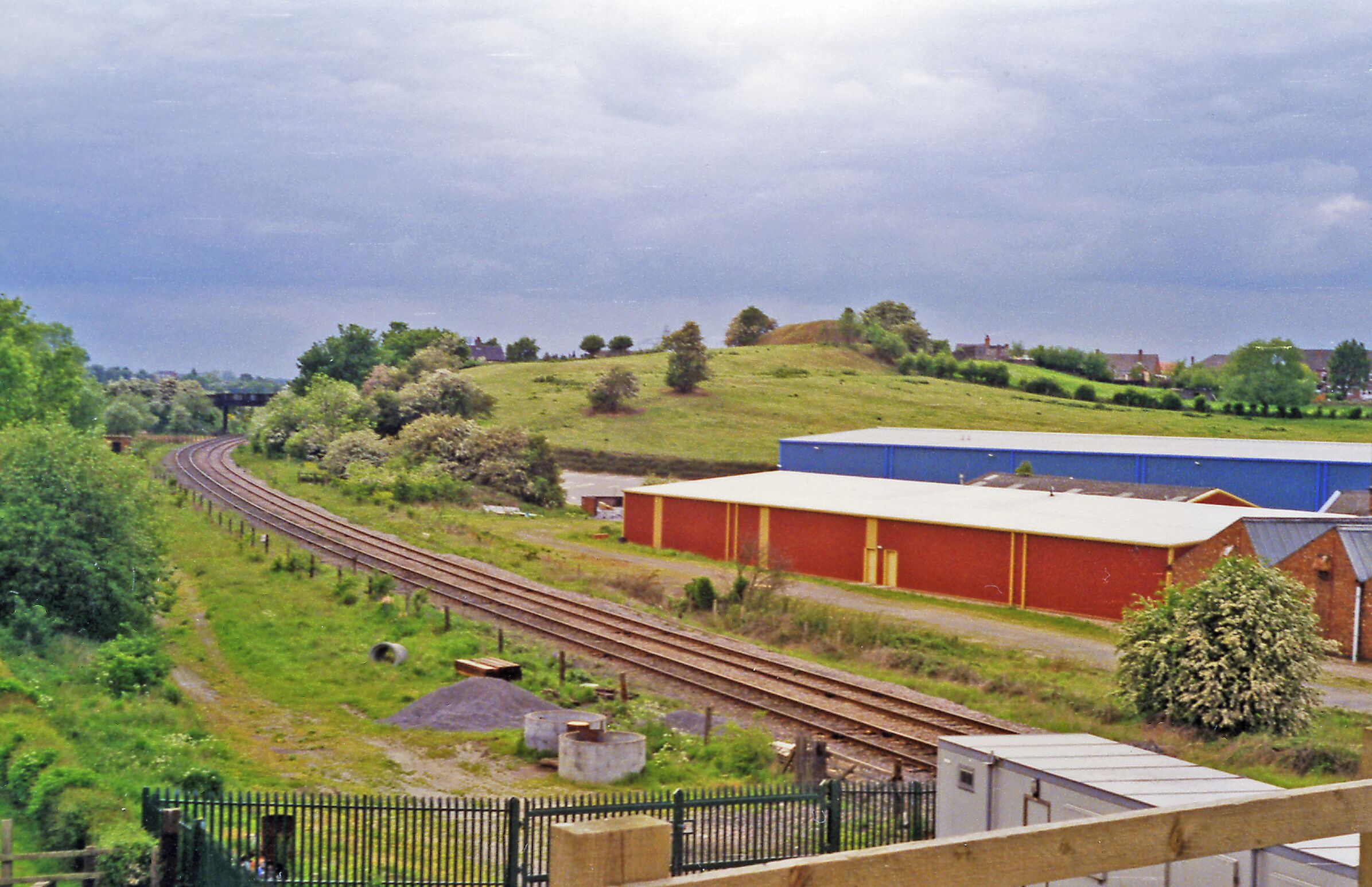 A railway track curving through a green landscape with a red building and a blue structure in the background.