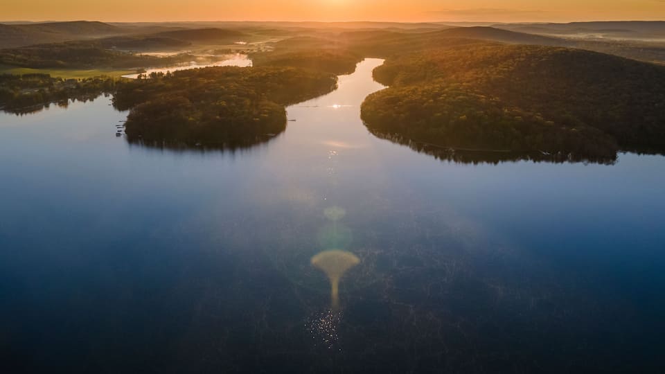 A serene landscape with a river reflecting the sun, surrounded by forested hills.