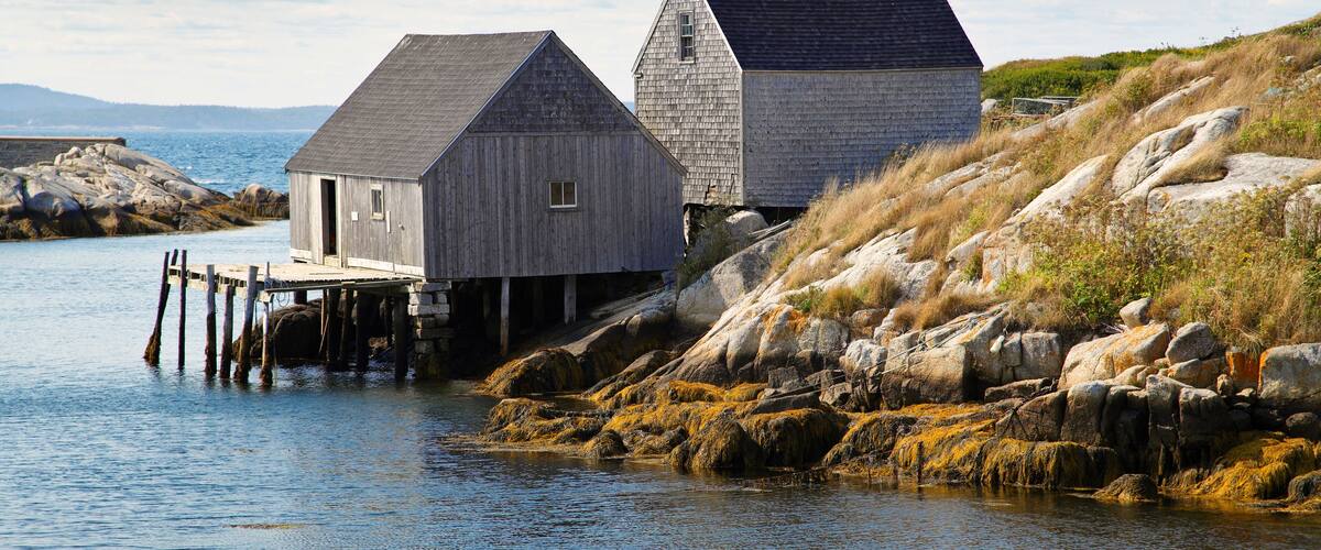 Two wooden buildings by a rocky shoreline with a calm body of water.