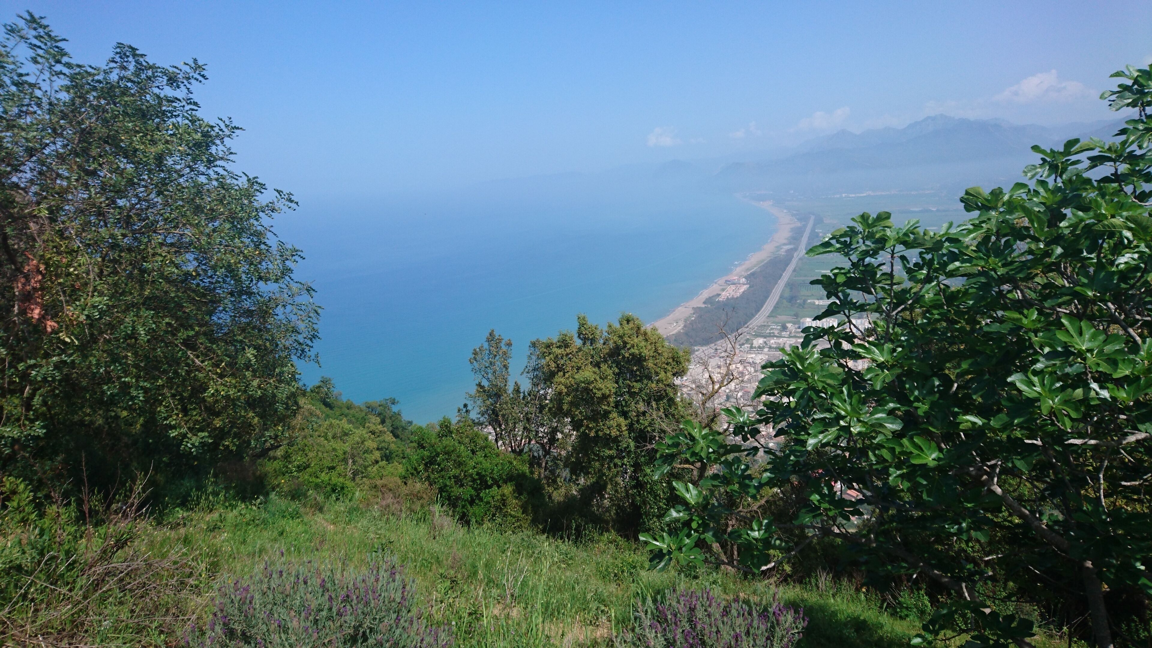 A coastal landscape with a clear view of the sea, a beach, and a city in the distance.