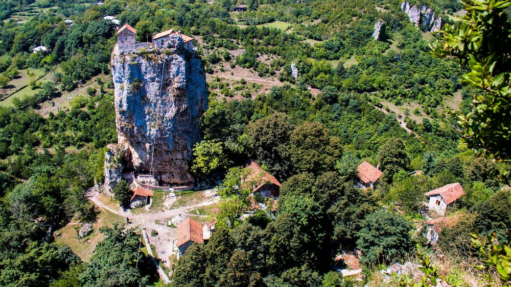 A rugged cliff with a small building on top, surrounded by dense greenery and scattered houses.