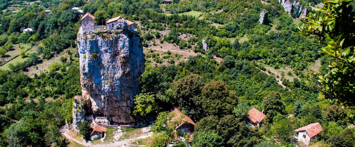 A rugged cliff with a small building on top, surrounded by dense greenery and scattered houses.