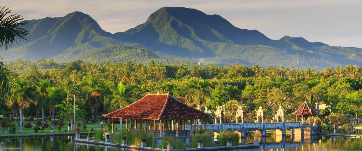 A serene lake with a traditional pavilion, lush greenery, and mountains in the background.