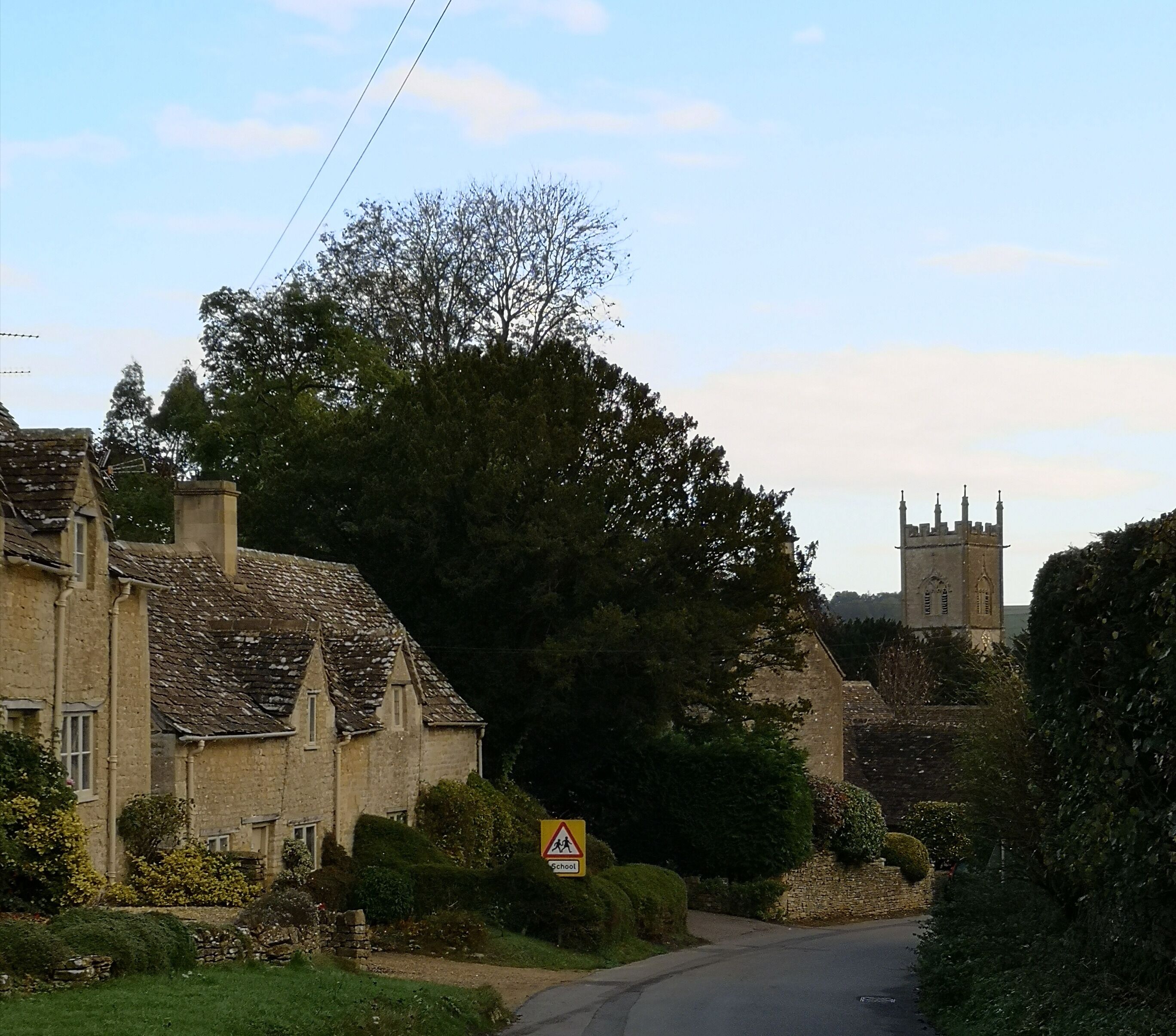 A rural road with stone houses, a school warning sign, and a church tower in the distance.