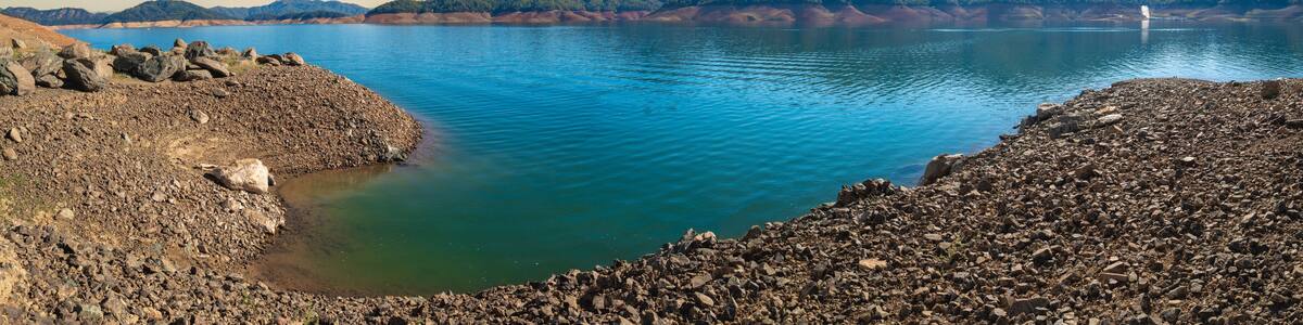 Shasta Lake at low water level with the gravel lake bed exposed in the Sacramento River, Northern California, United States, panoramic landscape with curving water’s edge