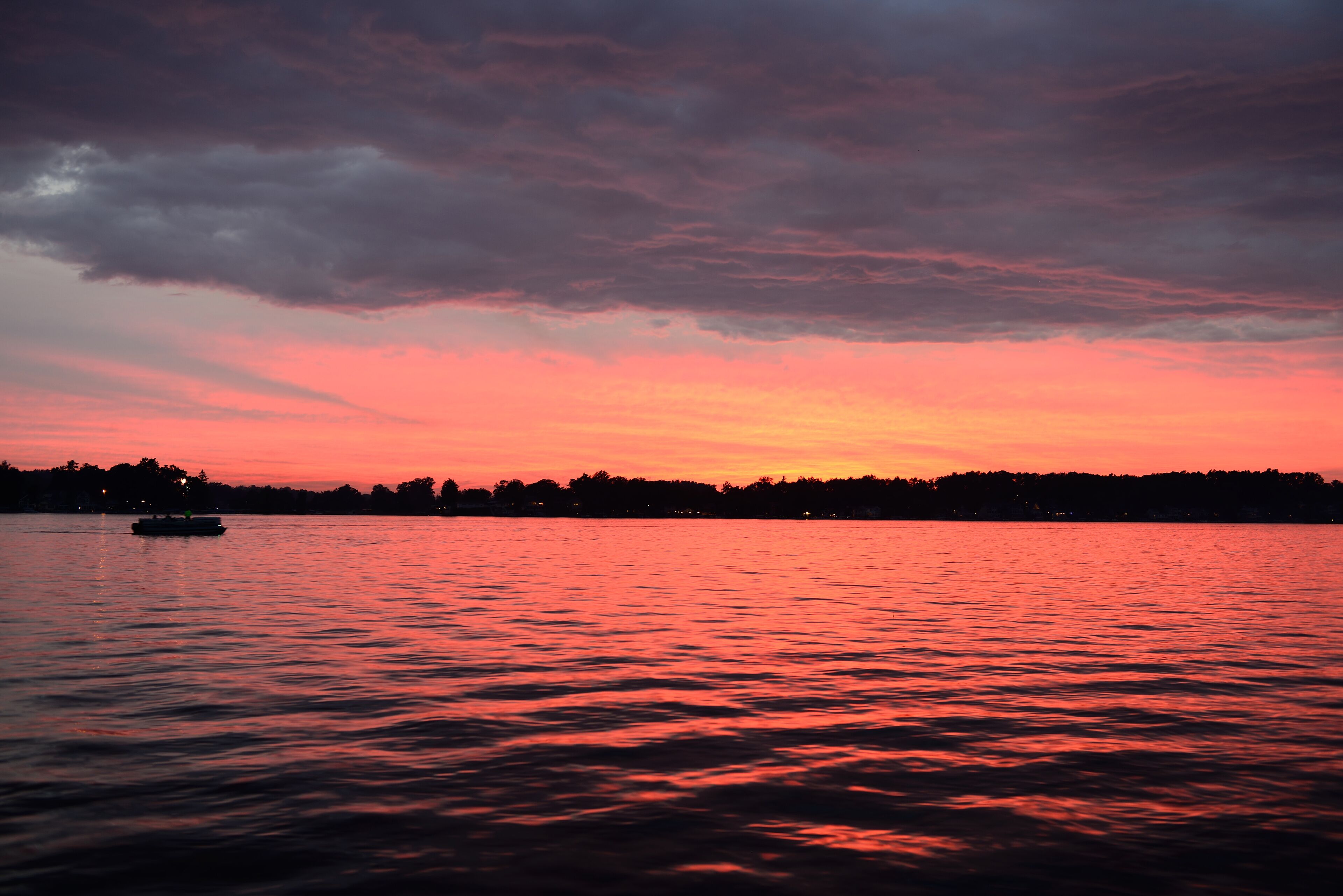 A sunset over a body of water with a boat and a forested shoreline.