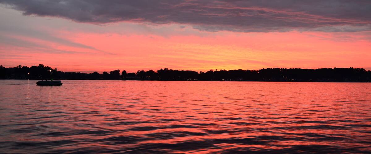 A sunset over a body of water with a boat and a forested shoreline.