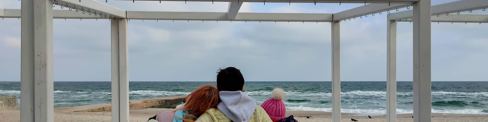 Three people in warm jackets and hats sitting closely on a white wooden bench, embracing and gazing at the waves of a cloudy sea, creating a cozy and emotional winter beach scene