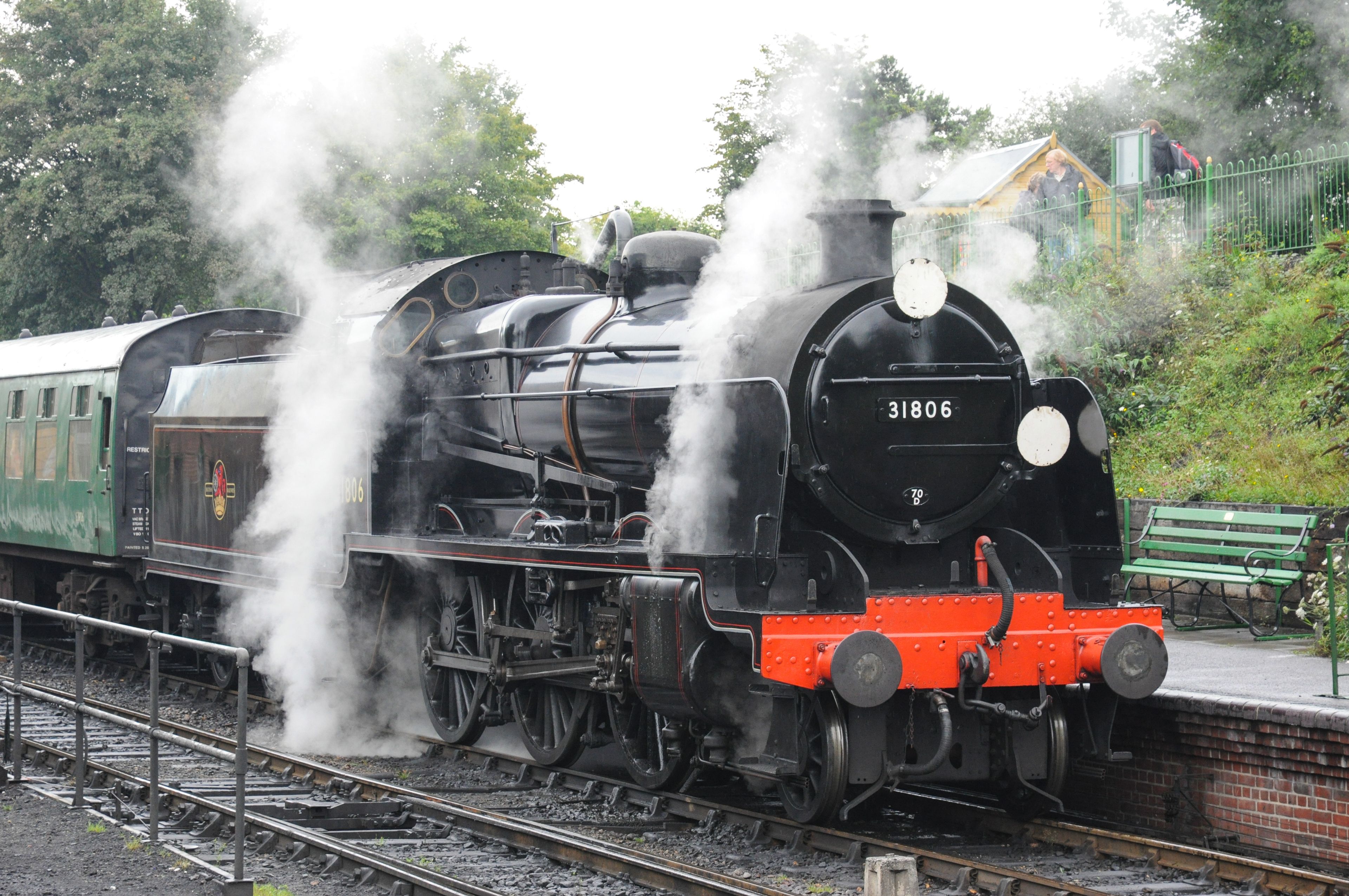 Una locomotora de vapor con el número 31806 sobre una vía férrea, expulsando vapor, con un banco verde y una cerca al fondo.