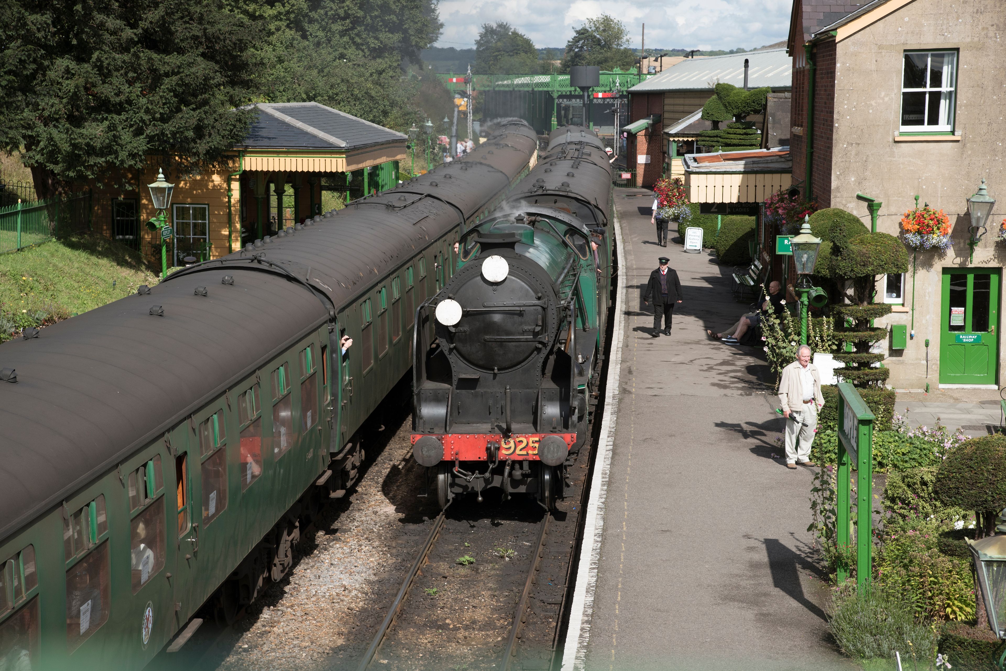Eine Dampflokomotive zieht grüne Waggons an einem Bahnhof mit einem grünen Gebäude und Blumenkörben.