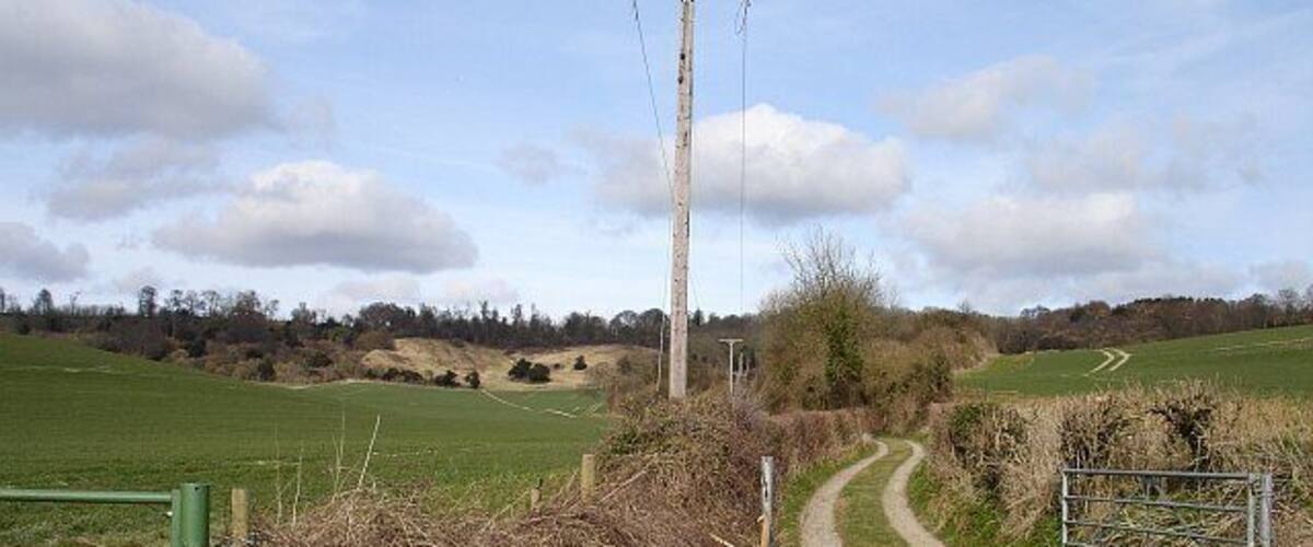 A rural dirt path leading to a gate, surrounded by fields and a utility pole.