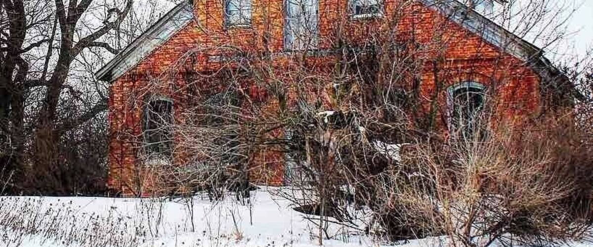 A red brick building with multiple windows and a chimney, surrounded by bare trees and snow.