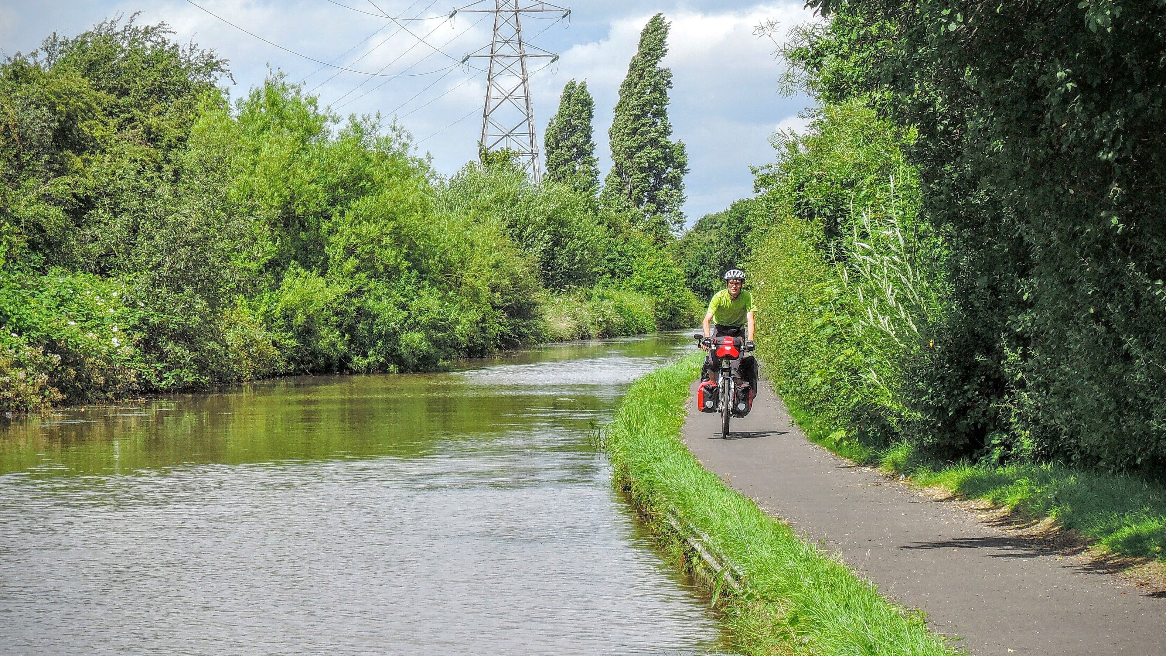 A cyclist on a paved path alongside a river, surrounded by lush greenery and tall trees.