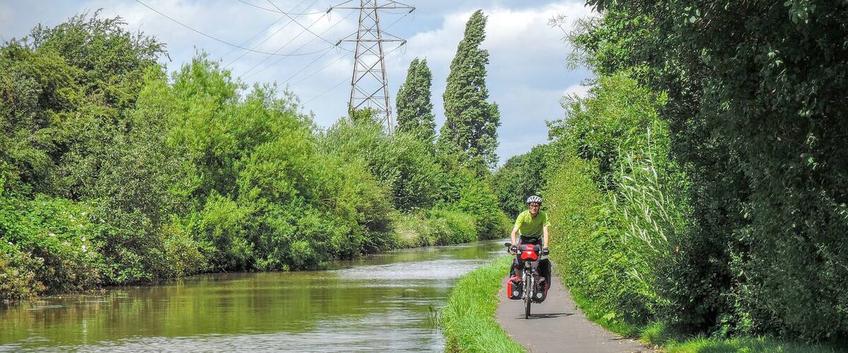 A cyclist on a paved path alongside a river, surrounded by lush greenery and tall trees.