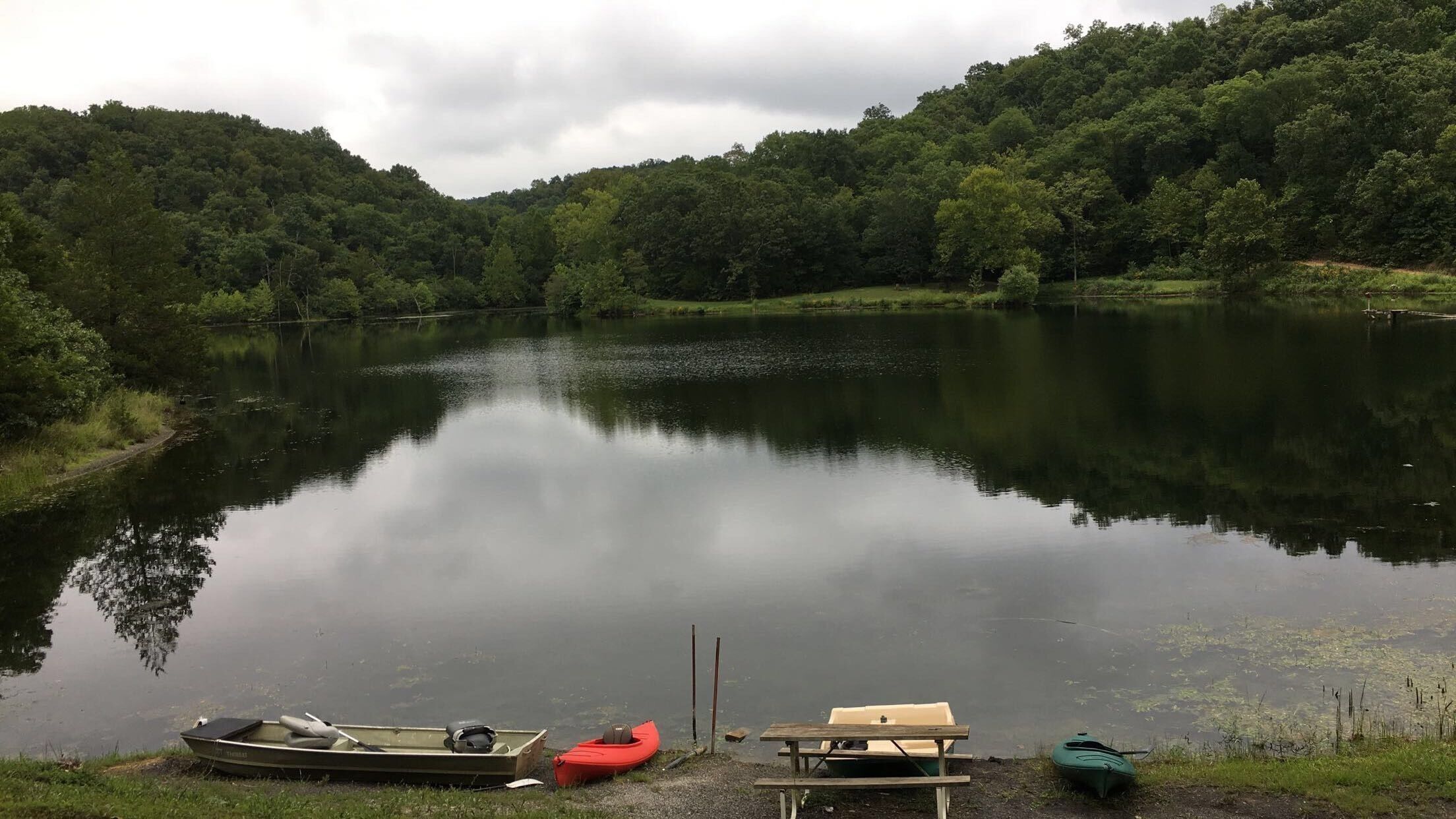 A calm lake surrounded by dense forest, with boats and a picnic table on the shore.
