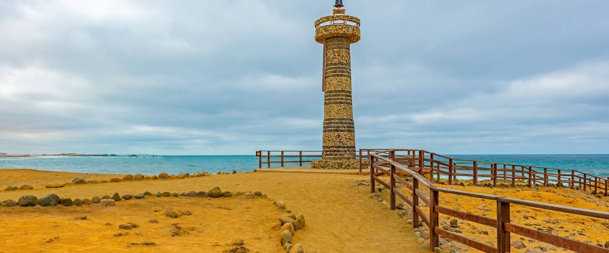 A coastal lighthouse with a wooden walkway leading to it, surrounded by a sandy beach and the ocean.