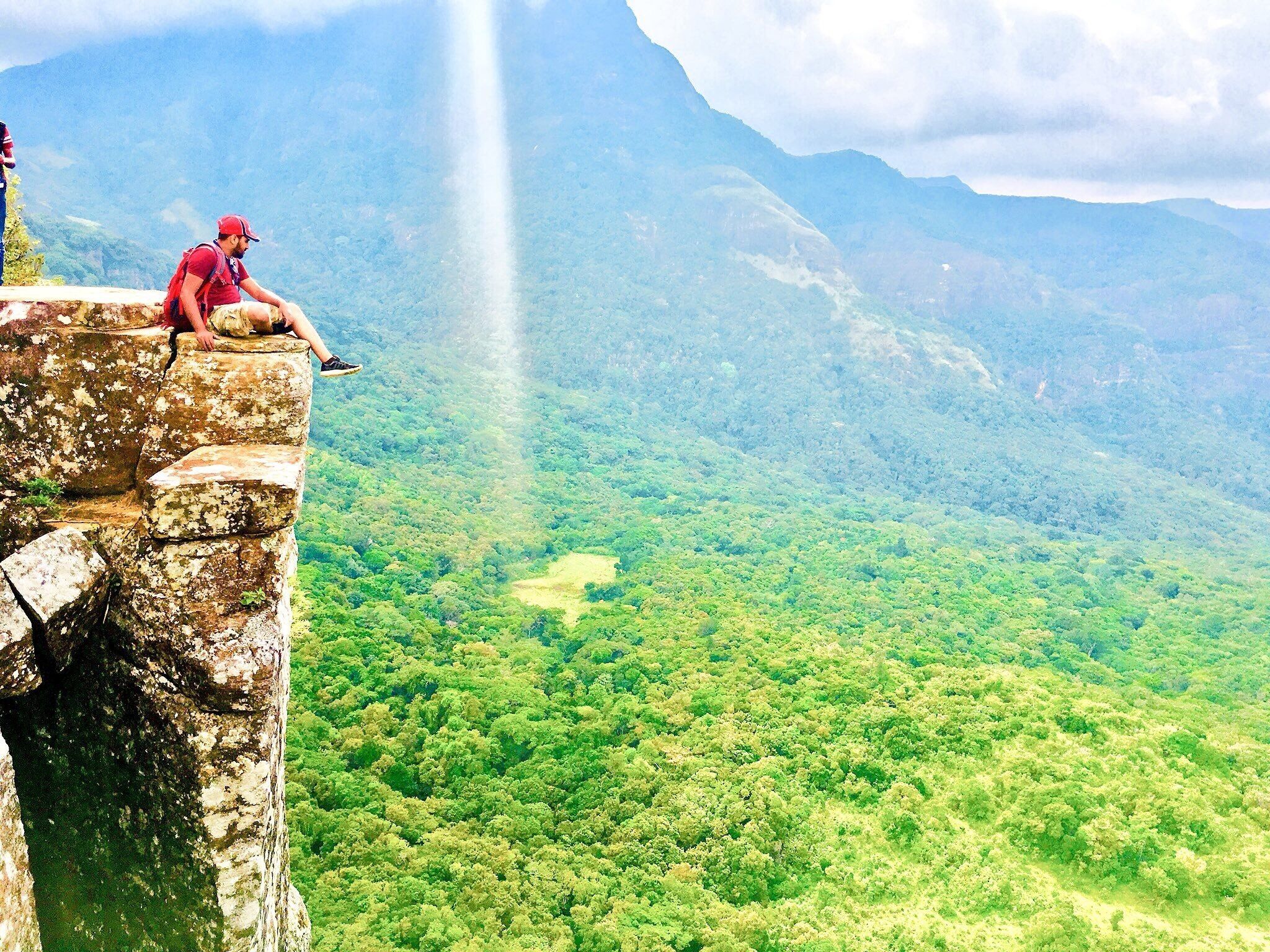 A person sitting on a cliff edge overlooking a lush green valley with mountains in the background.