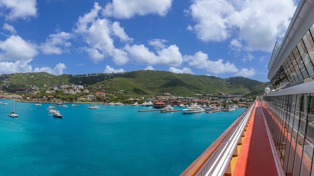Ein Schiffsdeck einer Kreuzfahrtschiffs mit Blick auf einen Hafen, der voller Boote und Yachten ist, umgeben von einer gebirgigen Küstenlinie.