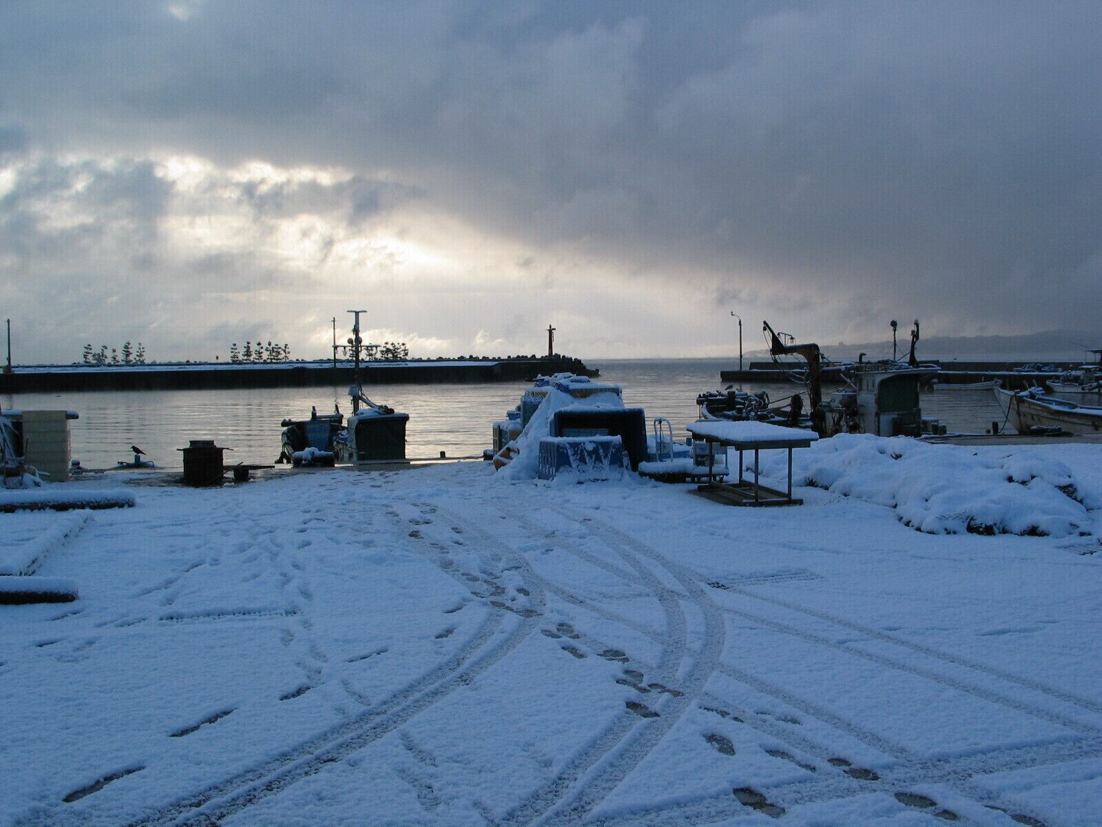 A snowy waterfront with boats docked, a snow-covered table, and footprints in the snow.