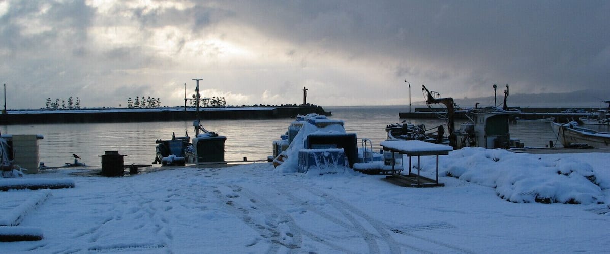 A snowy waterfront with boats docked, a snow-covered table, and footprints in the snow.