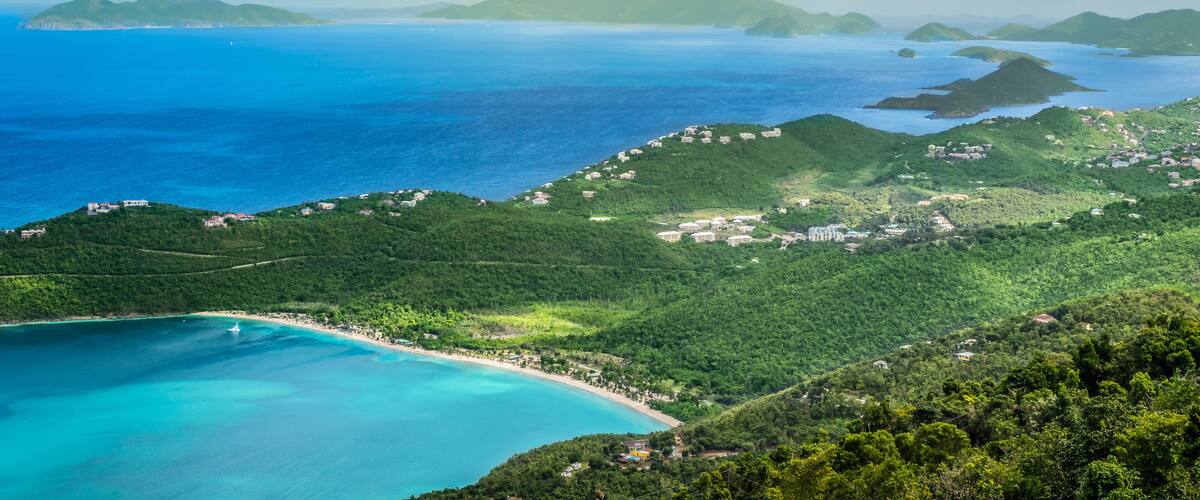 A coastal landscape with a beach, lush green hills, and a clear blue sea.