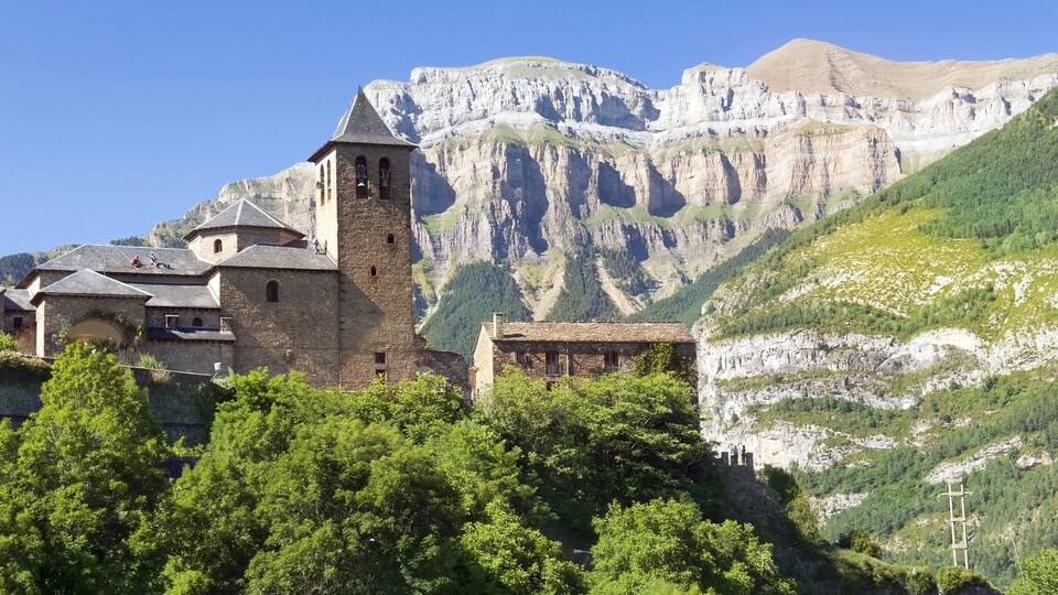 A historic stone church with a bell tower, set against a backdrop of rugged mountains and lush greenery.