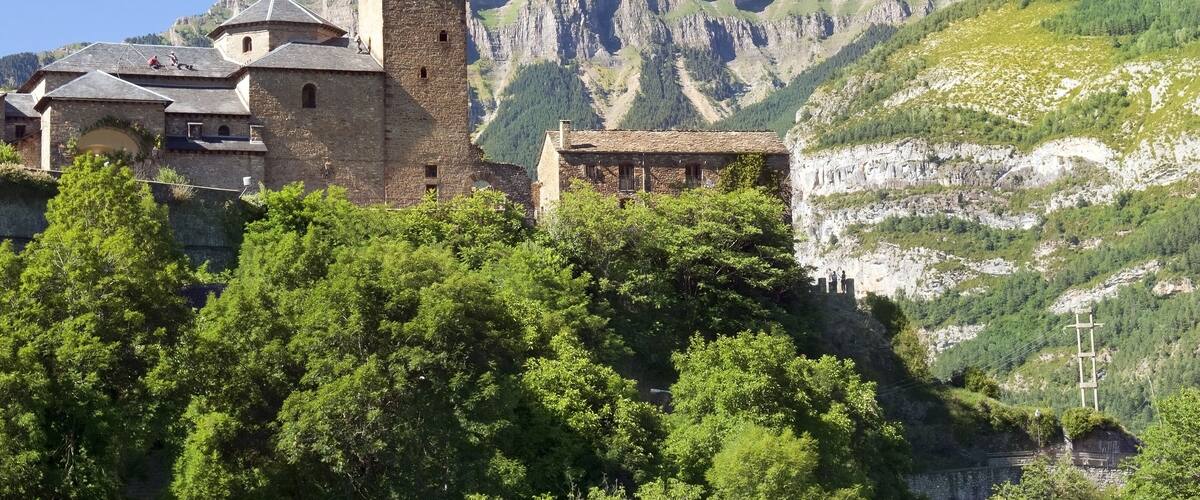 A historic stone church with a bell tower, set against a backdrop of rugged mountains and lush greenery.