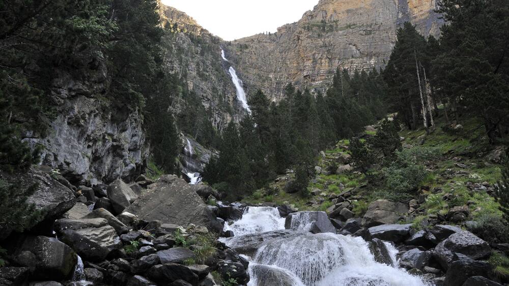 Une cascade d’eau se déversant sur des rochers dans une vallée boisée, avec une montagne en arrière-plan.