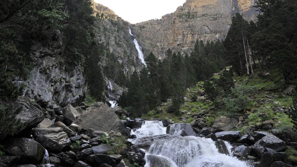 A waterfall cascading over rocks in a forested valley with a mountain in the background.