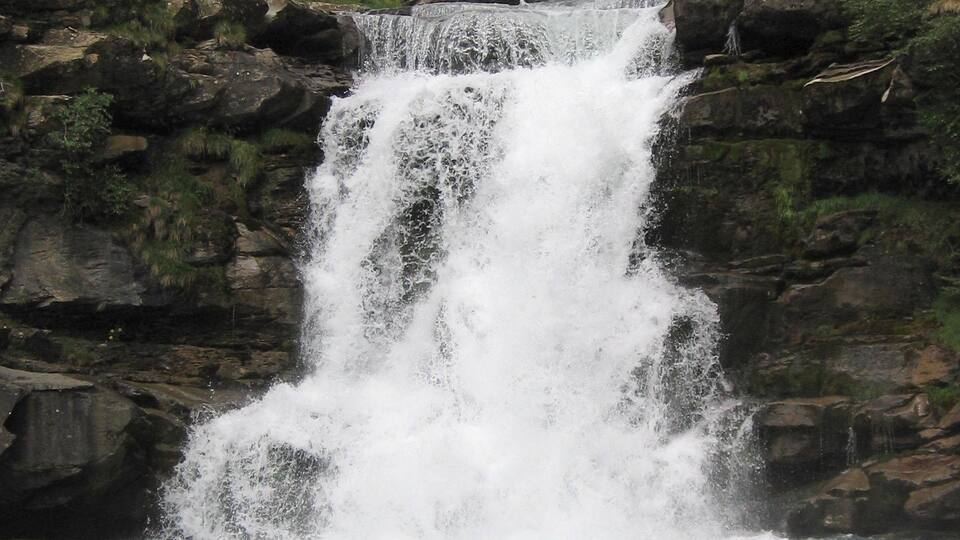 A waterfall cascading over rocks into a river surrounded by forested hills.