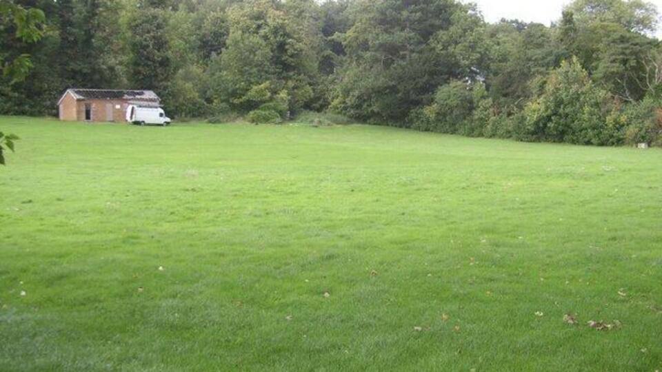 A grassy field with a small building and a white vehicle in the distance, surrounded by dense trees.