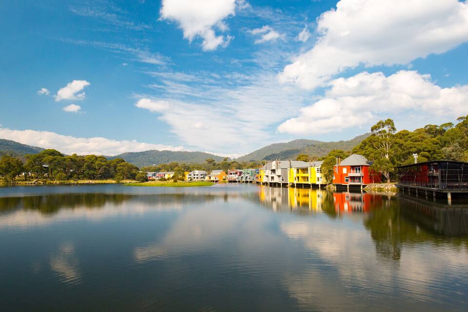 A calm lake with colorful houses reflecting on the water, surrounded by green hills and a clear blue sky.