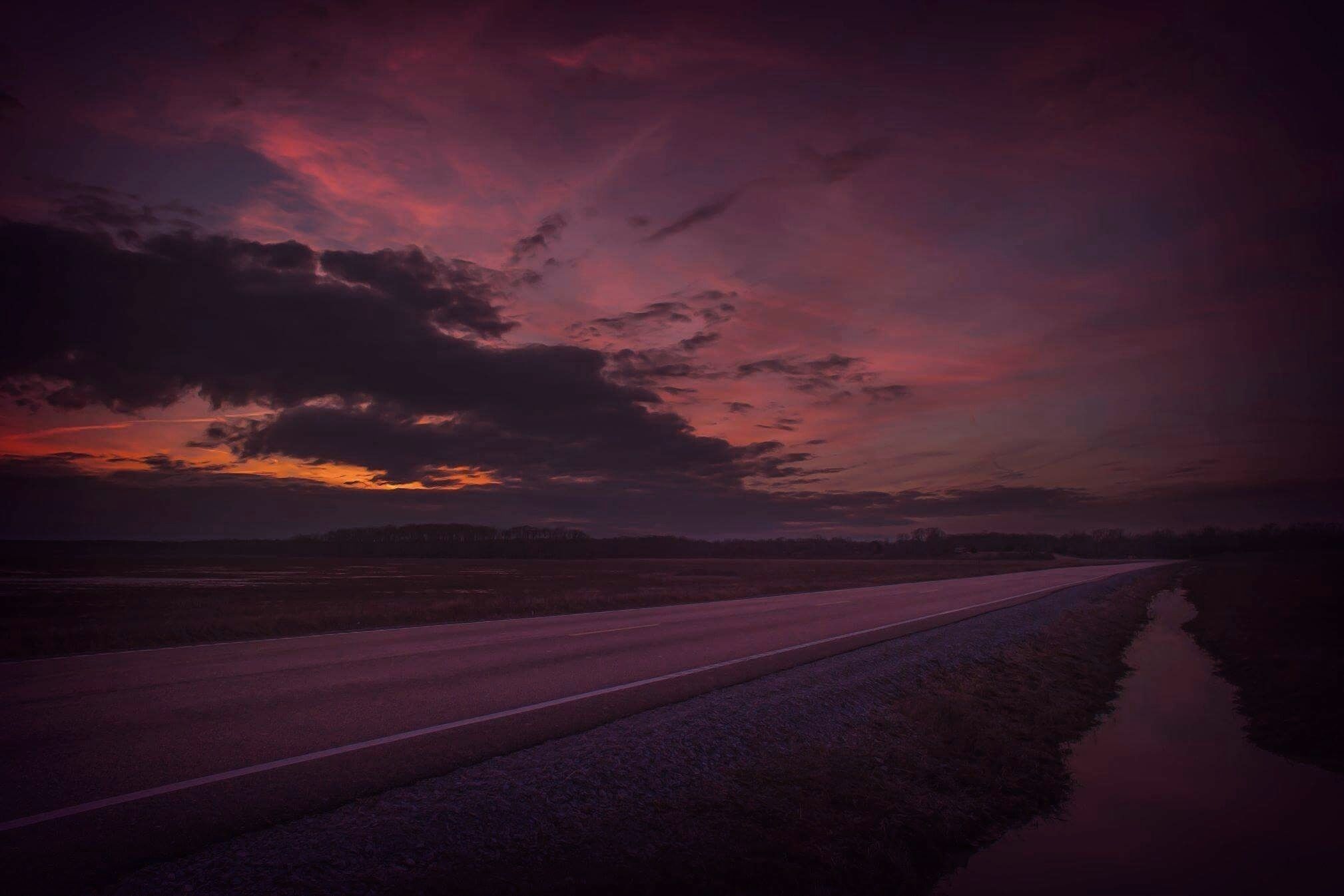 A road with a reflective water surface on the right, surrounded by fields under a dramatic sunset sky.