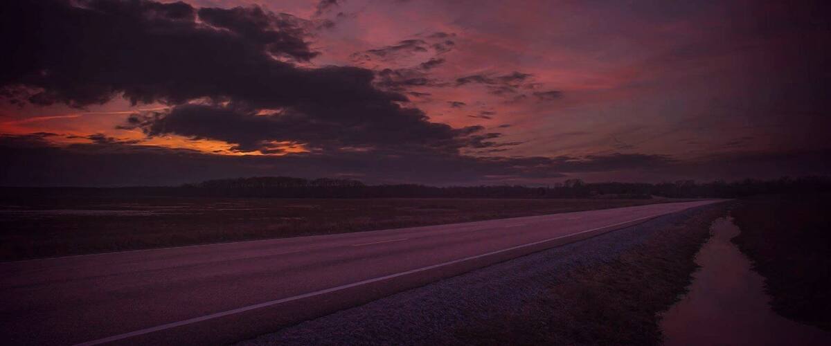 A road with a reflective water surface on the right, surrounded by fields under a dramatic sunset sky.