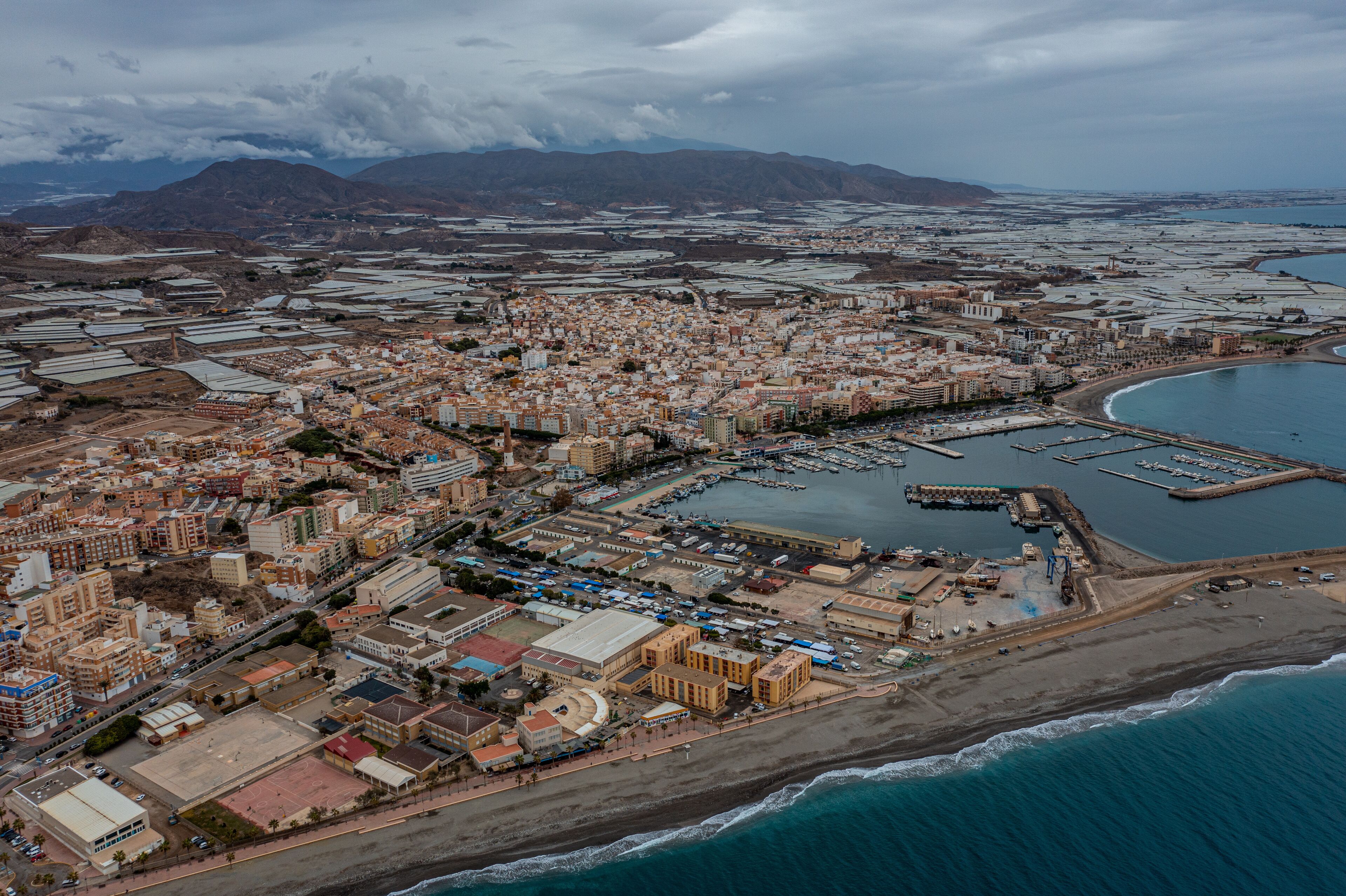 An aerial view of a coastal city with a marina, industrial areas, and mountainous background.