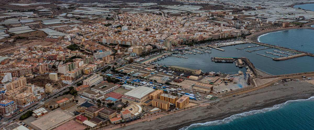 An aerial view of a coastal city with a marina, industrial areas, and mountainous background.
